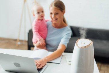 Selective focus of humidifier near woman using laptop and holding baby daughter clipart