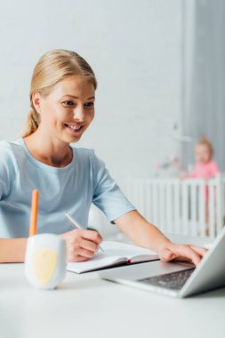 Laptop kullanan ve bebek monitörünün yanındaki not defterine yazı yazan seçici bir kadın. 