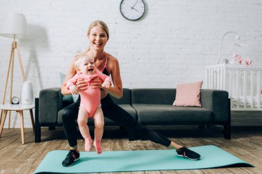 Sporcu kadın spor paspası üzerinde hamleler yapıyor ve bebeğini taşıyor. 