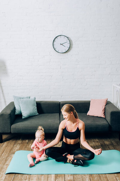 Mother in sportswear sitting in yoga pose near infant daughter on fitness mat