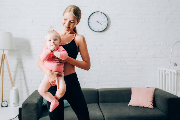 Fit woman in sportswear holding baby daughter on hands in living room 