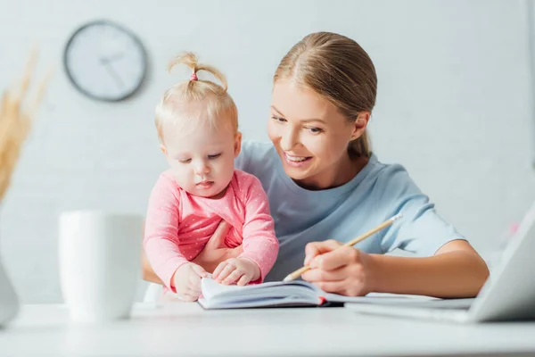 ノートに書く女性の選択的焦点とテーブルで作業しながら赤ちゃんの娘を保持 — ストック写真
