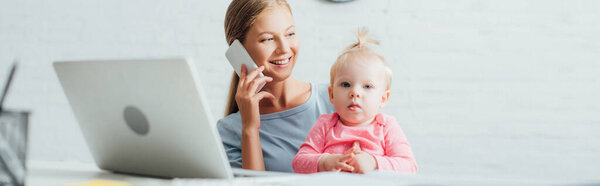 Panoramic shot of woman talking on cellphone and holding daughter near laptop on table 