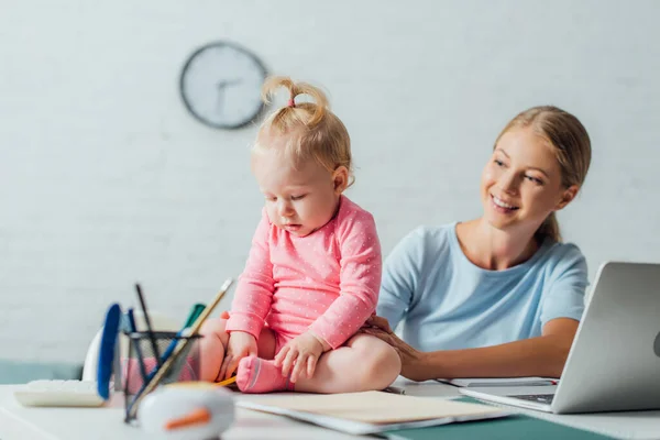 母親の横にあるテーブルの上に文房具やラップトップに座っている赤ちゃんの女の子の選択的な焦点 — ストック写真