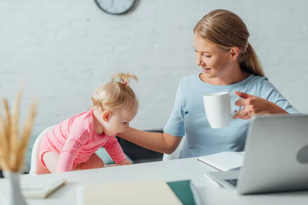 テーブルの上でカップを保持し ラップトップや文房具の近くに赤ちゃんの女の子に触れる女性の選択的な焦点 — ストック写真