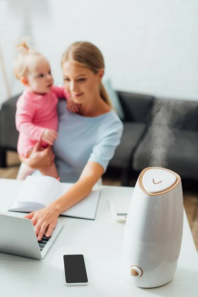 家庭でラップトップを使用している間 テーブルと母親の赤ちゃんの女の子を保持する加湿器の選択的な焦点 — ストック写真