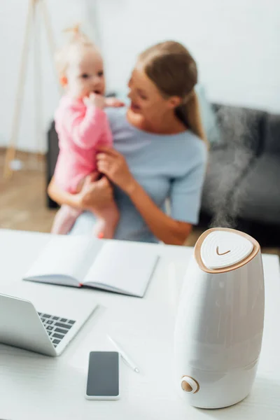 Selective focus of humidifier near gadgets on table and mother holding infant girl at background — Stock Image Selective Focus Humidifier Gadgets Table Mother Holding Infant Girl Background Royalty Free Stock Images
