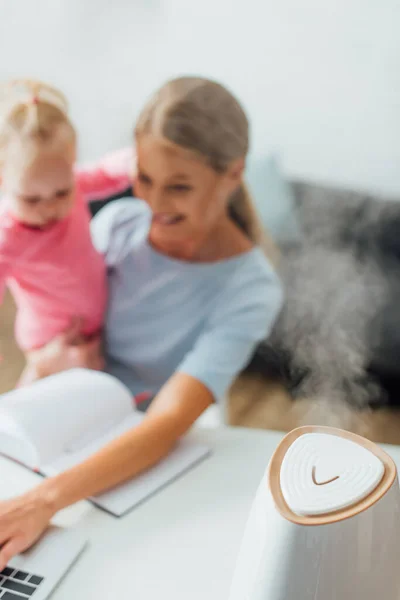 Selective focus of humidifier with mother holding baby girl and using laptop on background — Stock Image Selective Focus Humidifier Mother Holding Baby Girl Using Laptop Background Stock Image