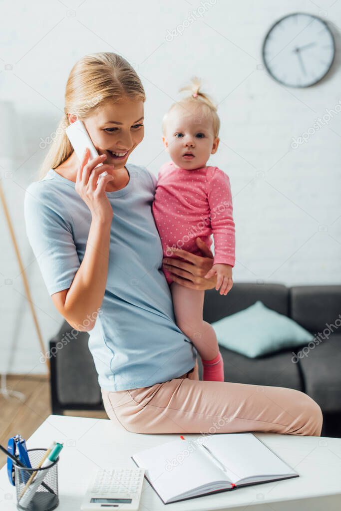 Selective focus of woman talking on smartphone and holding baby girl near stationery on table 