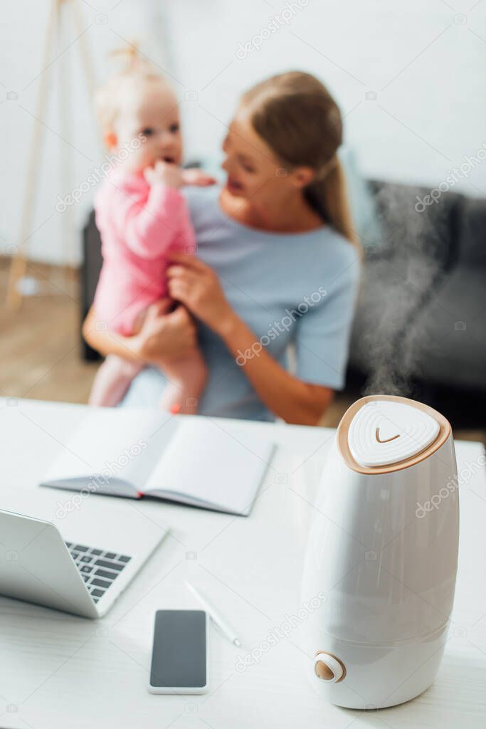Selective focus of humidifier near gadgets on table and mother holding infant girl at background 