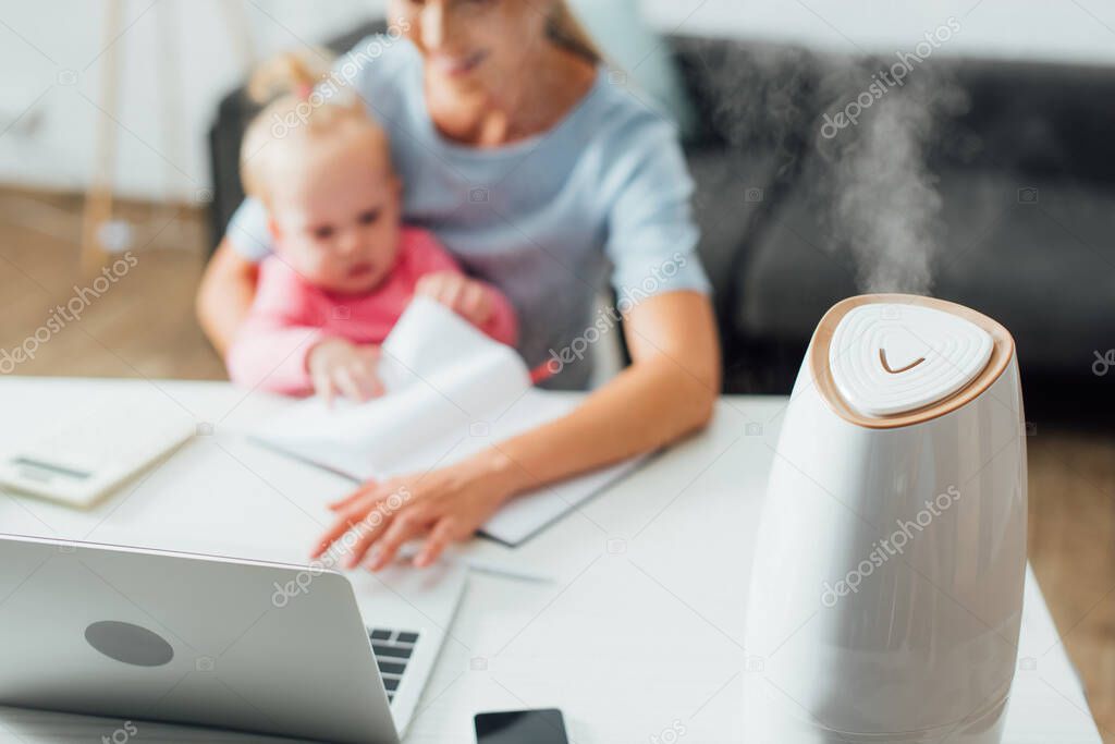 Selective focus of humidifier on table near woman holding baby girl and working at home 