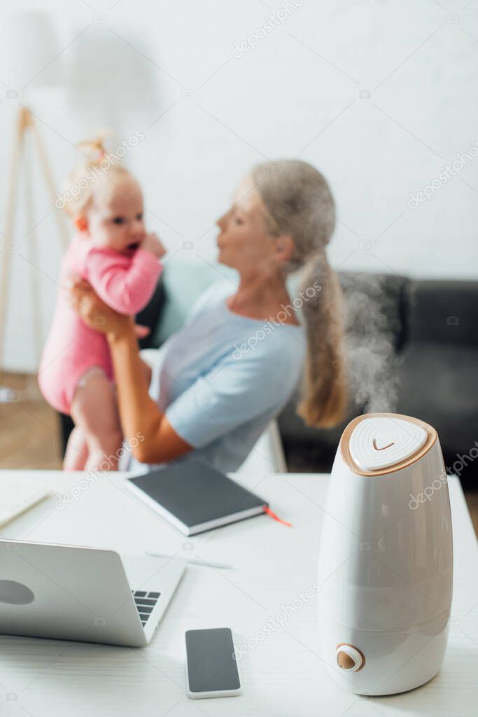 Selective focus of humidifier on table and freelancer holding baby girl at home 