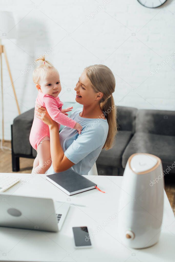 Selective focus of mother holding baby girl near gadgets, notebook and humidifier on table 