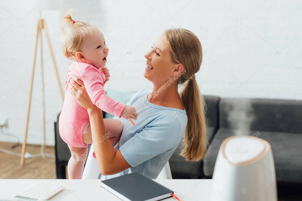 Selective focus of woman holding infant near notebook and humidifier on table 