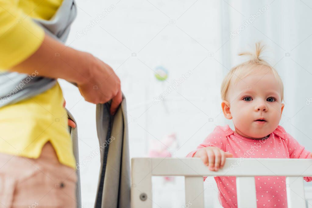 Selective focus of infant girl in crib looking at camera near mother in baby sling 