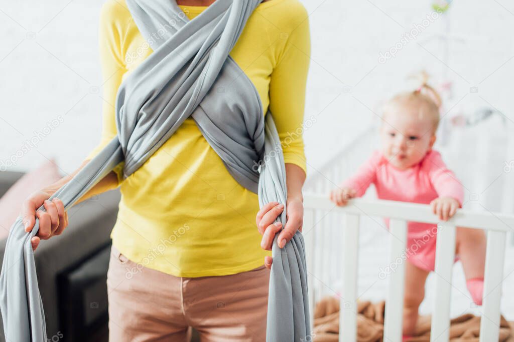 Selective focus of woman wearing baby sling near daughter in crib 