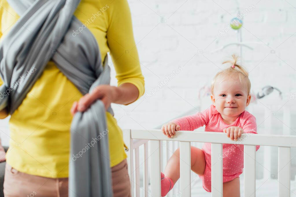 Selective focus of infant in baby crib near woman wearing sling at home 
