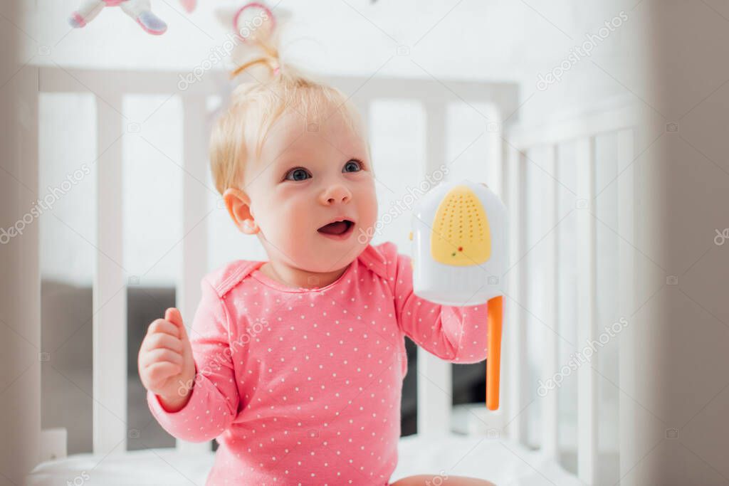 Selective focus of excited infant girl with baby monitor looking away in crib 