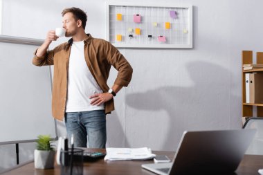 Genç işadamı ofiste dikilirken kahve içiyor gözleri kapalı ve eli kalçasında