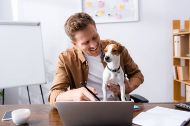 Genç iş adamı, Jack Russell Terrier ile çalışırken dizüstü bilgisayarını işaret ediyor.