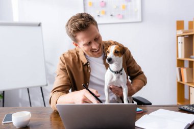 young businessman pointing with finger at laptop while working with jack russell terrier dog in office clipart