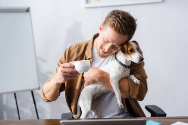 İş yerinde Jack Russell Terrier 'a sarılırken elinde kahve fincanı tutan genç bir iş adamı.