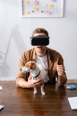 VR kulaklıklı genç işadamı Jack Rassell Terrier köpeğinin yanında iş yerinde.