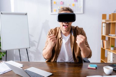 Heyecanlı iş adamı, iş yerinde VR kulaklığı kullanırken kazanan hareketi gösteriyor