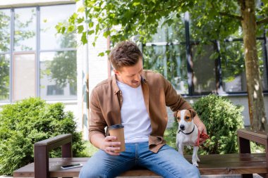 Jack Russell Terrier köpeği ve kahve ile bankta oturan genç adam yeşil bitkilerin yanına gitmek için