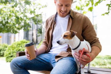 Günlük kıyafetli bir adam Jack Russell Terrier 'ın yanında bankta oturuyor ve elinde kahve tutuyor.
