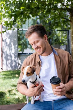 Jack Russell Terrier köpeği olan genç adam dışarıda otururken tek kullanımlık kupa tutuyor.