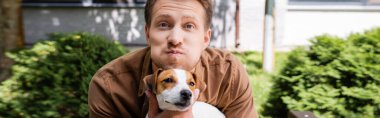Jack Russell Terrier köpeği tutan, yüzünü buruşturan ve yanaklarını şişiren adamın yatay konsepti