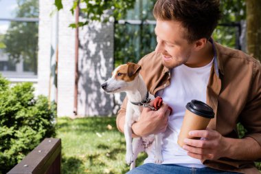 Jack Russell Terrier 'ı tutan genç adam elinde kahveyle bankta otururken