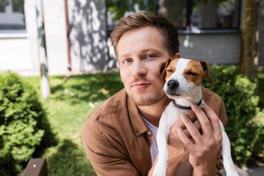 Genç adam dışarıda Jack Russell Terrier 'a sarılırken kameraya bakıyor.