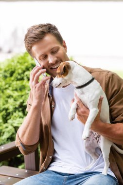 Günlük kıyafetli bir adam Jack Russell Terrier köpeğiyle bankta oturuyor ve akıllı telefondan konuşuyor.