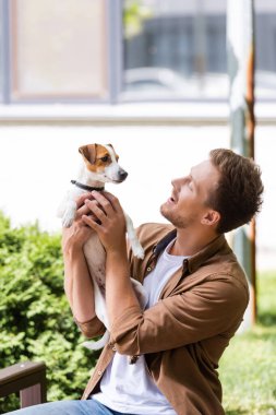 Dışarıda bankta otururken Jack Russell Terrier köpeğini tutan gündelik kıyafetli genç adam.