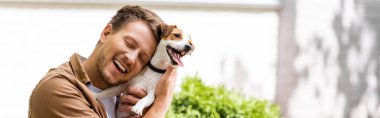 Kapalı gözlü bir adamın Jack Russell Terrier köpeğine sarılmasının panoramik konsepti 