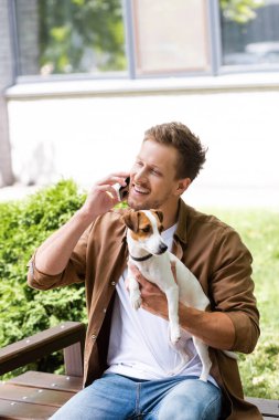 Jack Russell Terrier 'ın köpeğini tutan ve bankta otururken telefonla konuşan gündelik kıyafetli genç adam.