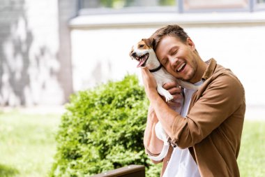 Gözleri kapalı genç adam yeşil çalıların yanında otururken Jack Russell Terrier köpeğine sarılıyor.