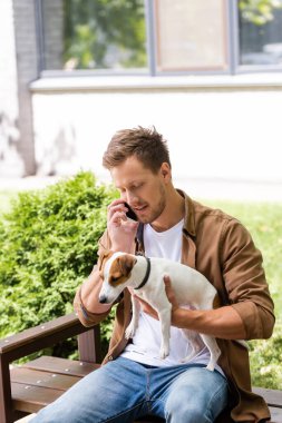 Gündelik giysiler içindeki genç adam akıllı telefondan konuşuyor ve bankta otururken Jack Russell Terrier köpeğini tutuyor.