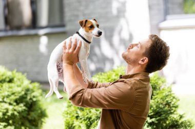 Genç adam dışarıda duruyor ve Jack Russell Terrier köpeği yetiştiriyor.