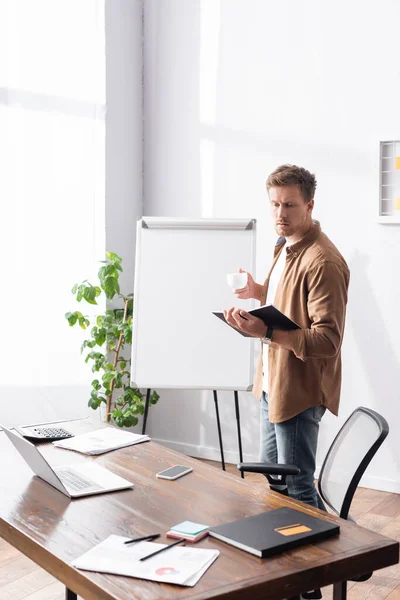 Young Businessman Holding Cup Coffee Notebook While Standing Flipchart Office — Stock Photo, Image