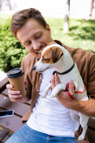 young man in casual clothes holding paper cup and jack russell terrier dog while sitting on bench