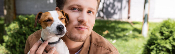 panoramic shot of man cuddling jack russell terrier dog while looking at camera