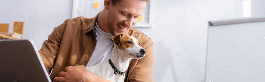 horizontal crop of young businessman holding white jack russell terrier dog with brown spots on head in office