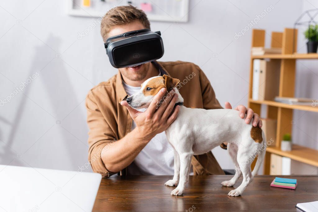 young businessman in vr headset touching jack russell terrier dog standing on office desk