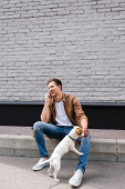Young man talking on smartphone near jack russell terrier on leash on urban street 