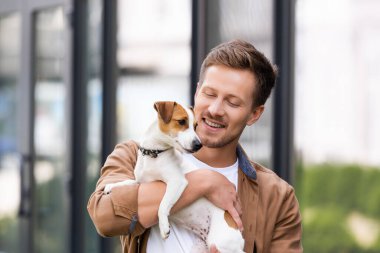 Genç adam beyaz Jack Russell Terrier köpeği tutuyor. Kafasında kahverengi spor var.