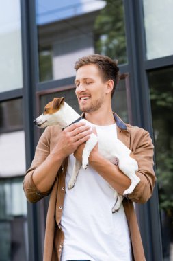 Günlük giysiler içinde genç bir adam Jack Russell Terrier köpeğini şehir sokaklarında tutuyor.