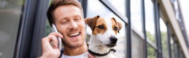 Jack Russell Terrier köpeğiyle sokakta akıllı telefondan konuşan genç adamın yatay görüntüsü.